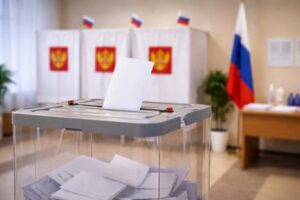 На фото запечатлён фрагмент избирательного участка. В центре кадра — прозрачная урна для бюллетеней с серой крышкой: в щель как раз опускают белый лист бумаги, а внутри уже лежит несколько сложенных бюллетеней. На заднем плане видны три кабинки для голосования — они задрапированы белой тканью с гербом Российской Федерации (двуглавый орёл на красном фоне). Над кабинками закреплены небольшие флажки с триколором. Справа стоит полноразмерный государственный флаг России, рядом — стол с документами и бутылками воды, а также комнатное растение в белом кашпо. Освещение мягкое, интерьер выдержан в нейтральных бежево‑белых тонах.