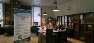 На фото — интерьер библиотечного. В центре — трое людей (две женщины и мужчина) за столами, перед ними ноутбуки и документы. Слева — вертикальный баннер с текстом и QR‑кодом. По периметру — высокие книжные шкафы с книгами и папками. На заднем плане — окна с жалюзи, над столами — подвесные светильники, на стене — круглые часы. В левой части — зона с креслами и комнатными растениями.