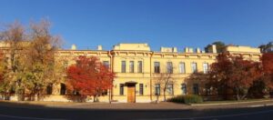 На фото — протяжённое трёхэтажное здание светло‑жёлтого цвета в стиле эклектики с классическими деталями: прямоугольными окнами, лепниной, декоративными парапетами. В центре фасада — входная группа с небольшим козырьком. По обе стороны от здания и перед ним — деревья с осенней листвой (от жёлто‑зелёной до ярко‑красной). На переднем плане — фрагмент асфальтированной дороги с закруглением, на фоне — чистое голубое небо.