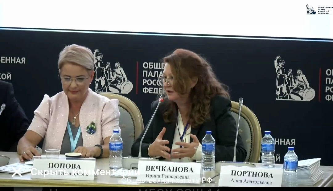 На фото — фрагмент заседания. За длинным столом сидят женщины в деловой одежде. Перед участницами — именные таблички, микрофоны, бутылки с водой, стаканы, стопки документов. Женщина слева в светло‑розовом пиджаке, с брошью, в очках, слегка улыбается, смотрит вниз. Женщина в центре в тёмном пиджаке, в очках, активно жестикулирует, говорит в микрофон. На тёмном фоне — белые надписи «Общественная палата Российской Федерации» и стилизованные силуэты людей. В правом верхнем углу — логотип Общественной палаты РФ