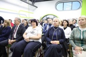 В зале на стульях рядами сидят участники мероприятия — преимущественно женщины и мужчины среднего и старшего возраста в деловой и полуделовой одежде. На переднем плане справа — женщина со светлыми волосами в светло‑зелёном жакете с узором и бежевых брюках; руки сложены на коленях, на левом запястье — часы. Рядом — женщина с короткими светлыми волосами в тёмно‑синем жакете с декоративной отделкой и тёмно‑синей водолазке; руки сложены на коленях. Левее — женщина с тёмными волосами в белой футболке с принтом и тёмных брюках; руки скрещены на груди. Рядом — мужчина с седыми волосами в тёмном костюме, белой рубашке и галстуке в диагональную полоску; руки сложены перед собой. На заднем плане — другие участники, часть из них смотрит вперёд, часть — в сторону. Интерьер современный: стены белые с лаймовыми акцентами и абстрактными линиями, вверху — овальное окно, видны элементы потолочной конструкции тёмного цвета. Освещение яркое, равномерное.