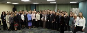 На фото — групповое фото большого коллектива в помещении, предположительно по случаю официального мероприятия. Люди стоят в несколько рядов: в центре — преимущественно мужчины и женщины в деловой одежде (костюмы, пиджаки, платья, блузки), многие носят бейджи на ленте. В переднем ряду справа — молодая женщина в белой блузке и тёмных брюках, слегка улыбается. Интерьер: стены нейтральных тонов, на фоне — плотные светло‑зелёные шторы, справа — декоративная белая панель с ажурным узором. На полу — ковёр с мелким пёстрым узором. Внизу кадра — спинки двух серых офисных кресел. Освещение искусственное, от потолочных светильников; композиция кадра — фронтальная, фокус на группе людей.